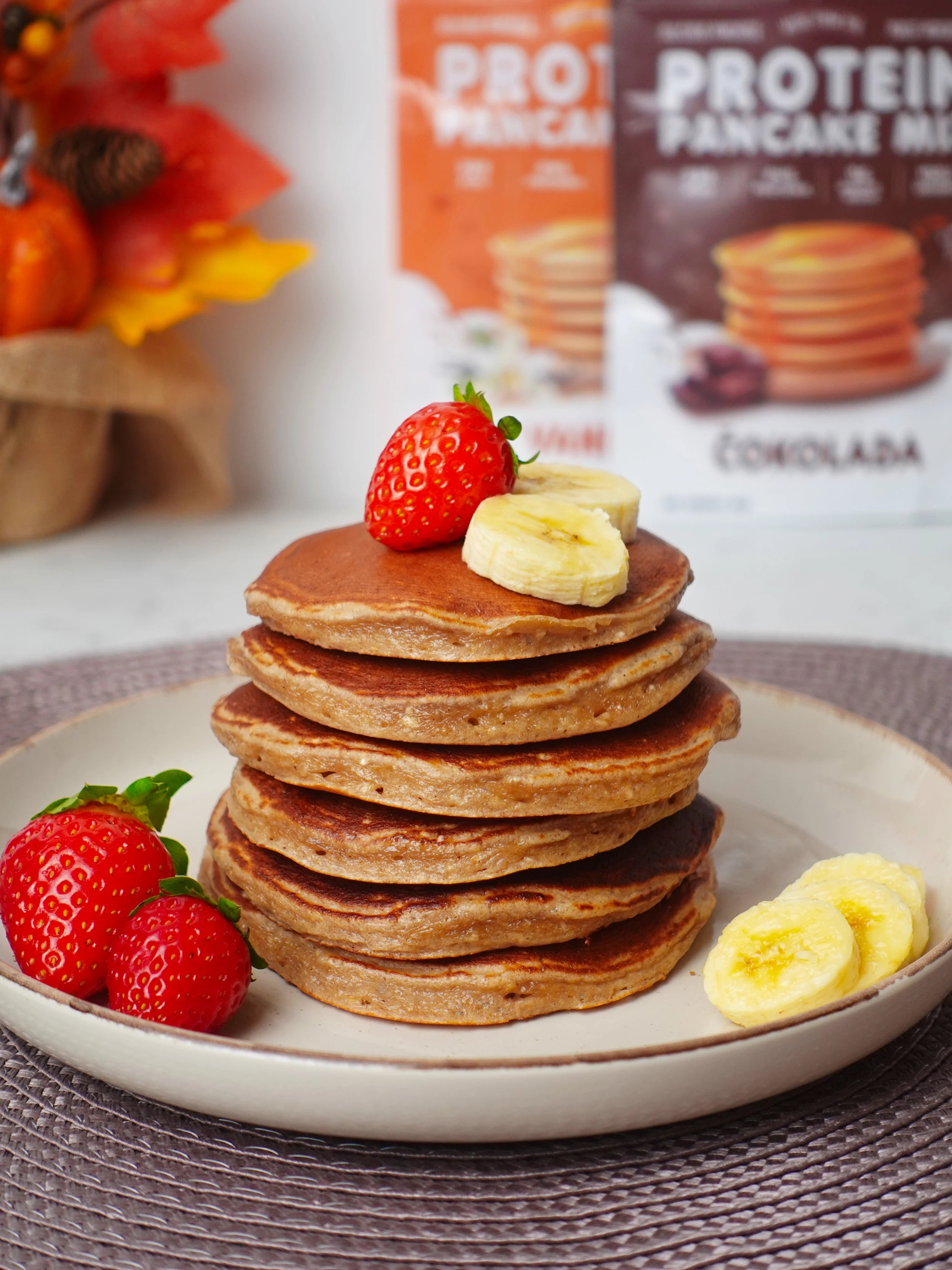 Proteinske palačinke sa jagodama i bananama na tanjiru, sa pakovanjem mix-a za proteinske palačinke u pozadini.